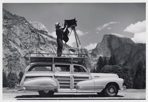 Ansel Adams standing on roof of Woody station wagon with large format camera, Half Dome behind him, Yosemite, circa 1942
