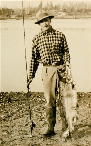 Eddie Bauer standing at riverbank holding a large salmon, plaid shirt and hat, circa 1930s